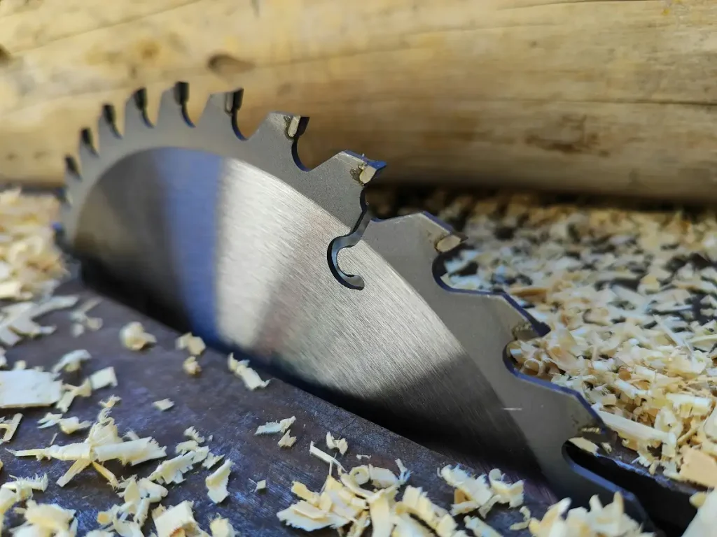 A close-up view of a metal circular saw blade with carbide teeth resting on a wooden surface, surrounded by fresh wood shavings and sawdust.