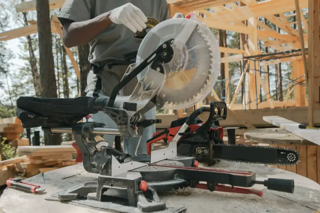 A miter saw and a red chainsaw sitting on a wooden surface at an outdoor cabin construction site, with a worker in the background adjusting a tool.